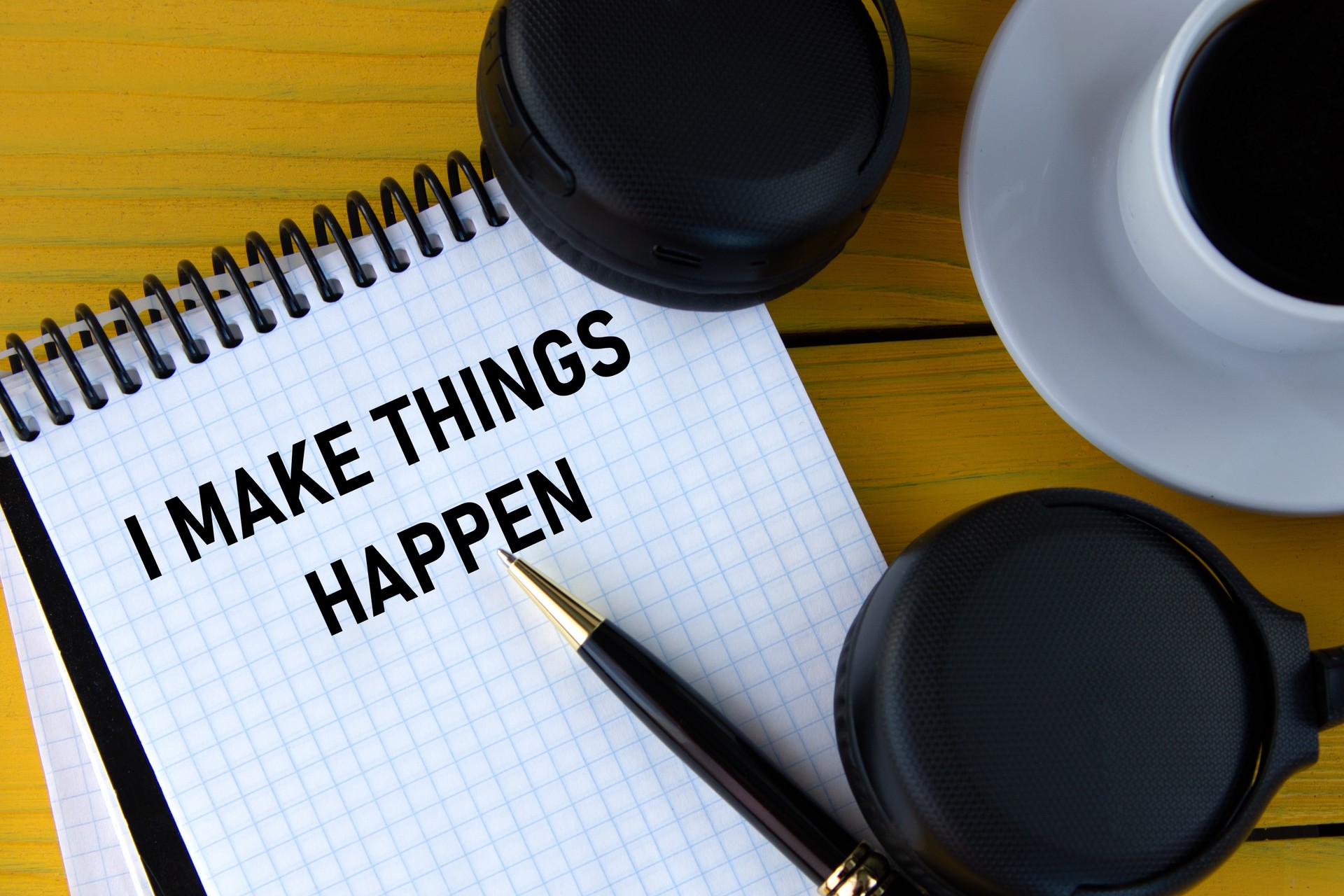 I MAKE THINGS HAPPEN - words on a white notepad on a yellow background with headphones, a cup of coffee and a pen